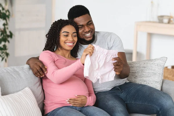 Sorrindo casal grávida preto segurando roupas de bebê — Fotografia de Stock