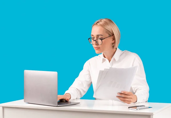 Focused female corporate employee with documents typing on laptop computer against blue studio background, free space