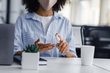 Covid salgını sırasında yeni normal ve virüs koruması. Koruyucu maskeli Afrikalı Amerikalı kadına odaklan. İş yerinde ellerini antiseptikle dezenfekte et. Dizüstü bilgisayarla.