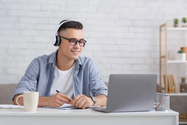 Σύγχρονη online εκπαίδευση. Χαμογελώντας τύπος με γυαλιά και ακουστικά βλέποντας σε απευθείας σύνδεση webinar ή το μάθημα κάνει σημείωση στο σημειωματάριο στο εσωτερικό του σαλονιού — Φωτογραφία Αρχείου