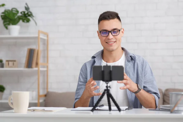 Βίντεο εγγραφής για το σύγχρονο blog ως ένα απομακρυσμένο έργο ή χόμπι. Ευτυχισμένος τύπος με γυαλιά κοιτάζει στο smartphone κάμερα σε τρίποδο, κάθεται στο tae στο σαλόνι — Φωτογραφία Αρχείου
