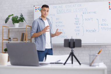 Salgın sırasında evde uzaktan eğitim. Genç adam öğrencilere İngilizce kurallarını açıklıyor, tahtaya yakın duruyor ve masadaki akıllı telefon webcam 'ine bakıyor.