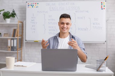 Uzaktan eğlenceli İngilizce dersleri ve koronavirüs salgını sırasında öğretmenlik. Öğrenciler için İngilizce kurallarını işaret eden ve açıklayan mutlu adam ve dizüstü bilgisayarın web kamerasına bakan