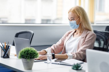 Yeni normal, modern ofis virüsten ve müdürlük işinden korunuyor. Koruyucu maskeli sarışın bir iş kadını elinde antiseptik şişesiyle bilgisayarda yazı yazıyor.