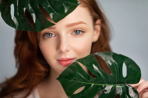 Mulher jovem de cabelos vermelhos posando segurando folhas tropicais sobre fundo cinza — Fotografia de Stock