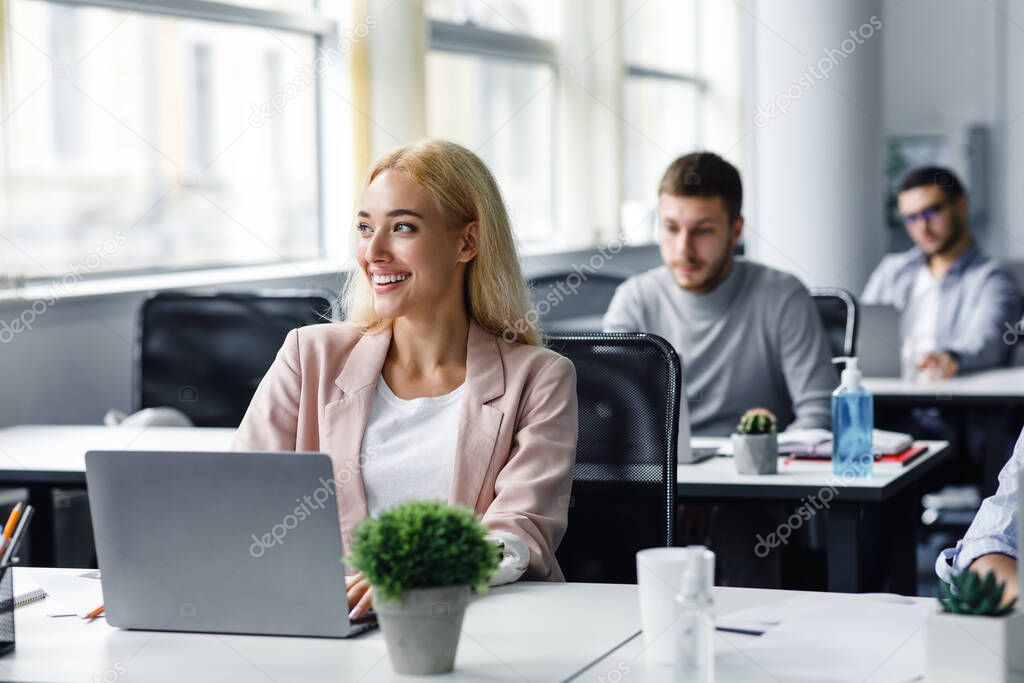 Work after lockdown. Happy young woman in suit looks out the window and works at laptop in office interior with plant in pot and antiseptics