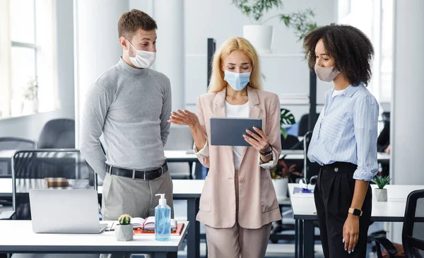 Vragen aan de manager tijdens het werk. vrouw baas in beschermende masker kijkt naar tablet en controles taak van guy en afrikaanse amerikaanse dame — Stockfoto