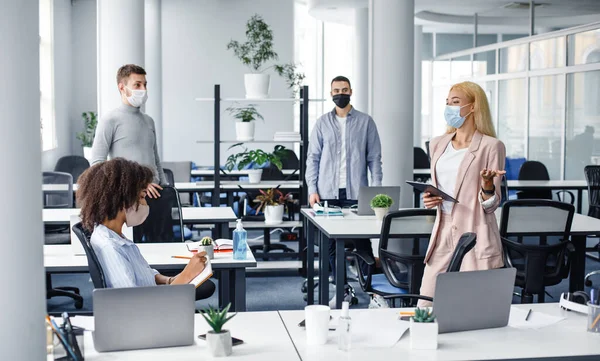 Vergadering in functie en instructies van de baas. Jonge zakenvrouw in beschermend masker met tablet in de hand spreekt tot werknemers op de werkplek — Stockfoto