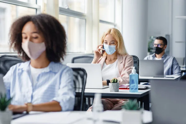 Pracownicy firmy pracują w biurze. Wielorasowi menedżerowie w maskach ochronnych w miejscach pracy z antyseptykami i laptopami, skupienie się na blondynce, rozmowa przez telefon — Zdjęcie stockowe