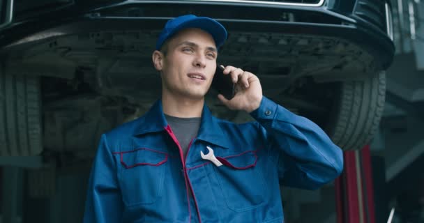 Bakım servisinde cep telefonuyla konuşan genç bir tamirci müşteriyle araba hasarını tartışıyor. — Stok video