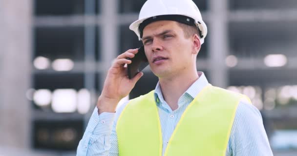 Σοβαρός επιστάτης σε προστατευτικό κράνος μιλάει με τον εργολάβο σε smartphone στέκεται στο εργοτάξιο — Αρχείο Βίντεο