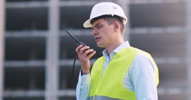 Foreman i hjelm og vest taler på walkie-talkie med bygherrer, stående på byggepladsen, tracking shot – Stock-video