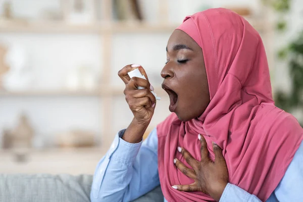 Чорношкіра мусульманка в хіджабі використовує інгалятор вдома, страждає дихальною проблемою — стокове фото