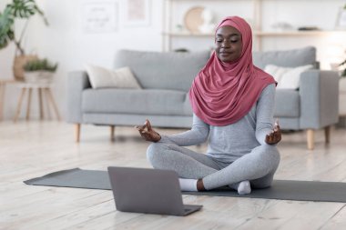Yoga devrede. Siyah Müslüman kadın evde dizüstü bilgisayarın önünde meditasyon yapıyor.