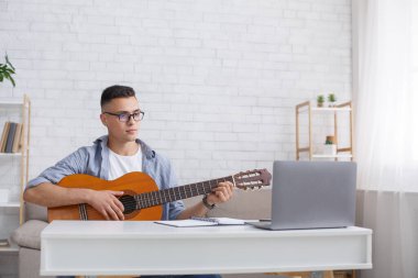 Modern teknoloji ve evde boş vakitlerde müzik öğrenmek için uygulama. Gözlüklü adam gitar çalıyor ve oturma odasının içinde dizüstü bilgisayara bakıyor.