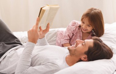 Genç baba küçük kızına kitap okuyor, yatağa uzanıyor.