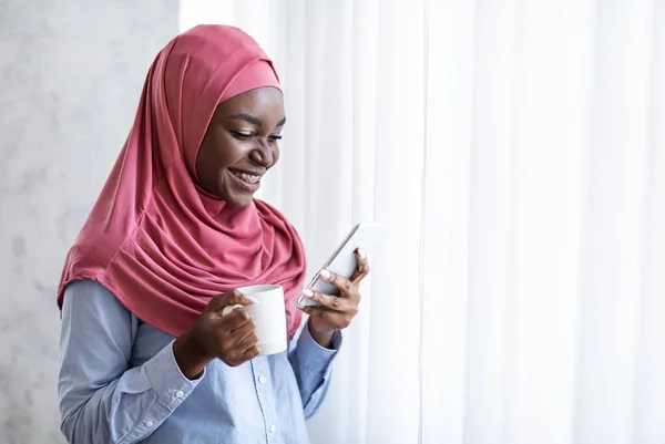 Relaxing Morning. Smiling Black Muslim Woman With Coffee And Smartphone Near Window — Stock Photo, Image