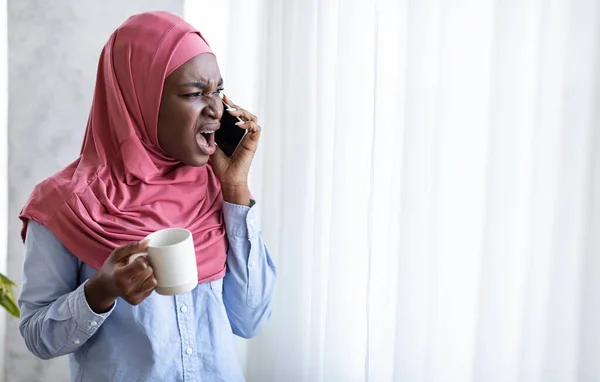 Nero musulmano donna litigando al telefono e bere caffè, in piedi vicino alla finestra — Foto Stock