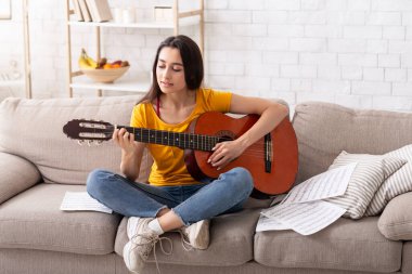 Evde hobilerin olsun. Güzel bir milenyum kadını oturma odasında kanepede akustik gitar çalıyor.