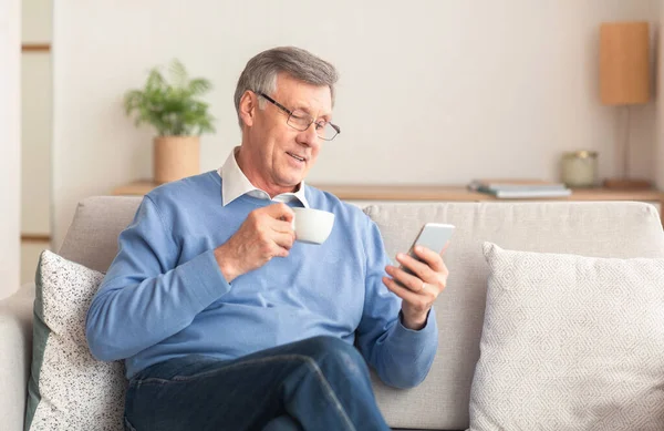 Homem sênior usando Smartphone tendo café sentado no sofá interior — Fotografia de Stock