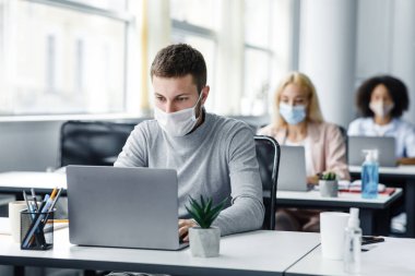 Coronavirus salgını sırasında birlikte çalışma ve güvenli çalışma. Yüz maskeli genç adam tecritten sonra işe geri döndü ve dizüstü bilgisayara baktı.