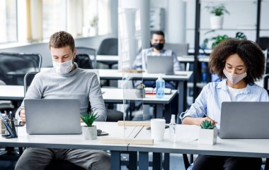 Yeni normal ve ofis işleri. Adam ve koruyucu maskeli Afro-Amerikan kadın iş yerinde antiseptik ve dizüstü bilgisayarlarla cam tahta üzerinde çalışıyorlar.