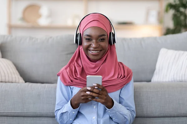 Wanita muslim kulit hitam menikmati musik di smartphone dengan headphone di rumah — Stok Foto