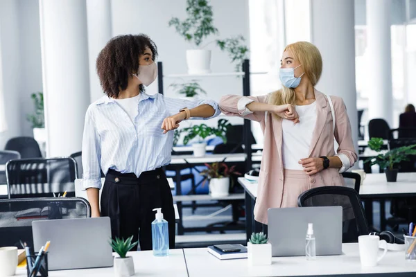 Nueva normalidad, protección contra el coronavirus y saludos durante la epidemia. Mujeres europeas y afroamericanas en máscaras protectoras saludan con sus codos — Foto de Stock