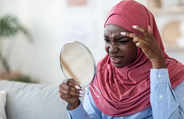 Проблемная кожа. Черная исламская женщина в хиджабе смотрит в зеркало дома — стоковое фото