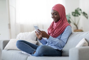 Evde boş zaman. Mutlu Afrikalı Müslüman Kadın Oturma odasında Akıllı Telefonla Dinleniyor