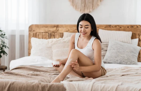 Jovem mulher asiática sentada na cama, aplicando loção corporal — Fotografia de Stock