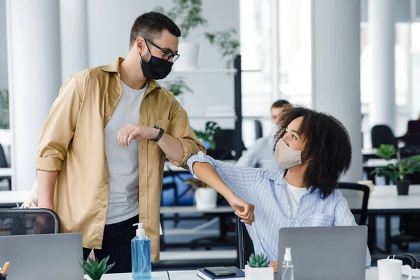 Hello colleague and new normal. Arabian guy and african american woman in protective masks greet with elbows at workplace in interior of office during epidemic