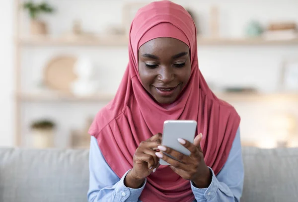 Mujer musulmana moderna en Hijab escribiendo mensaje en el teléfono inteligente en casa, relajarse en el sofá con gadget, enfoque selectivo, primer plano —  Fotos de Stock