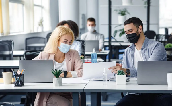 Social distance and work with documents in modern office. Millennial man and woman with papers in workplace with laptops and bottle of antiseptic talk after quarantine — Stock Photo, Image