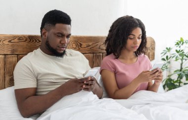 Siyah adam ve kadın yatakta oturuyor ve akıllı telefonlarını kullanıyorlar.