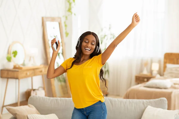 Happy African American lady with headphones and smartphone dancing to beautiful music at home
