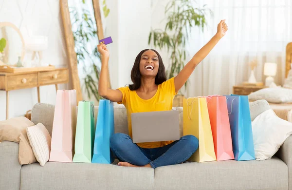 Excited black woman with credit card and laptop computer sitting on sofa surrounded by shopping bags, indoors
