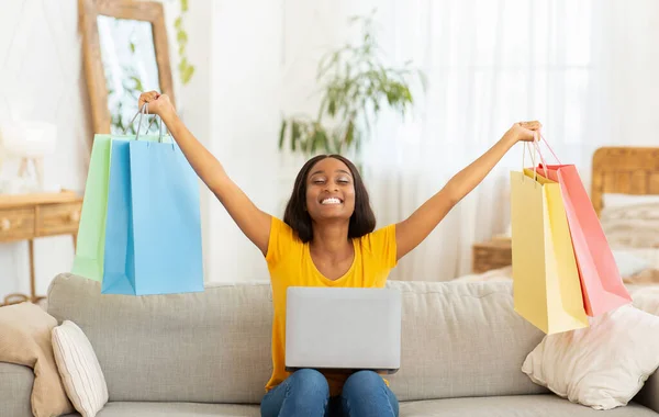 Excited black lady with laptop lifting hands full of shopping bags on sofa at home