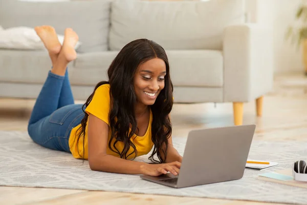Online life concept. Gorgeous black woman lying on floor with laptop computer, working or studying from home