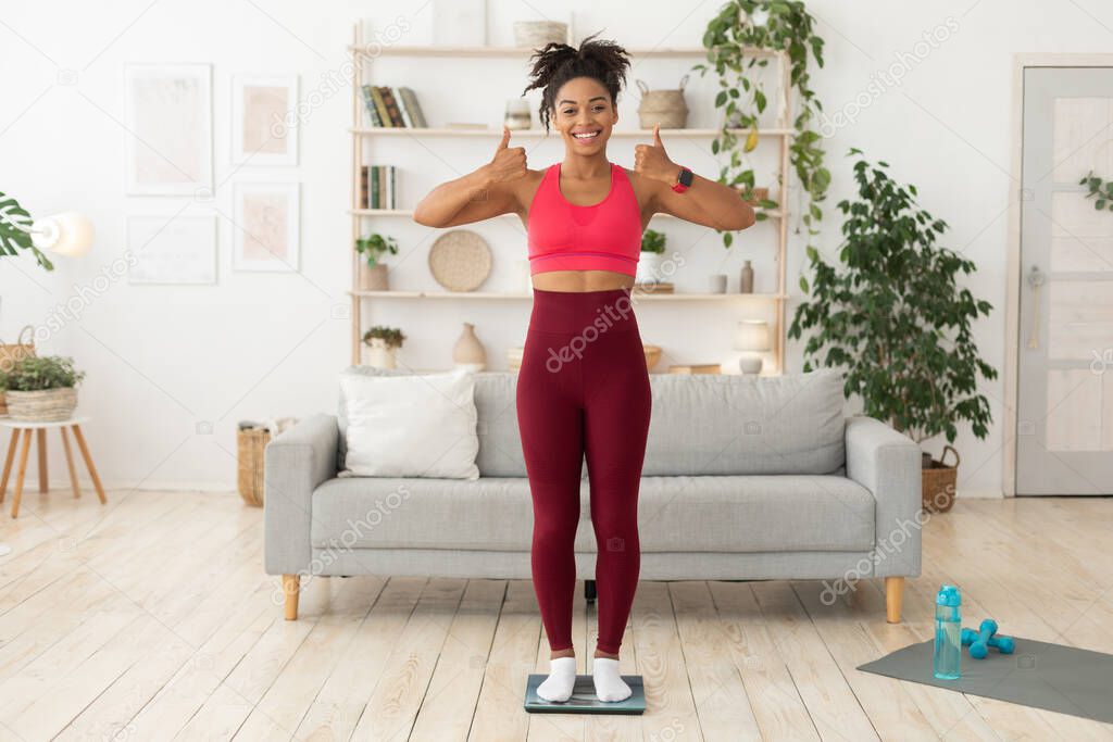 Black Lady After Weight-Loss Standing On Scales Gesturing Thumbs-Up Indoor