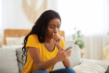 Endişeli siyahi kadın evde cep telefonundan iç karartıcı haberler okuyor.