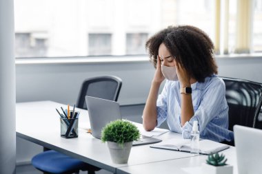 Aşırı çalışma, baş ağrısı ve hastalığın ilk belirtileri. Koruyucu maskeli Afrikalı kadın baş ağrısı çekiyor, masada dizüstü bilgisayar ve antiseptik ile oturuyor.