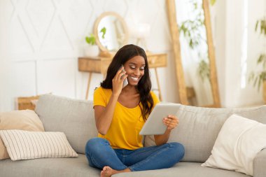 Siyah kadın evde akıllı telefonla konuşurken online iş, eğitim veya eğlence için tablet bilgisayar kullanıyor