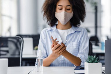 Coronavirüs salgını sırasında ofiste sağlığın korunması. Koruyucu maskeli genç Afrikalı Amerikalı kadın ellerini antiseptikle dezenfekte ediyor, iç kısımdaki masada oturuyor.