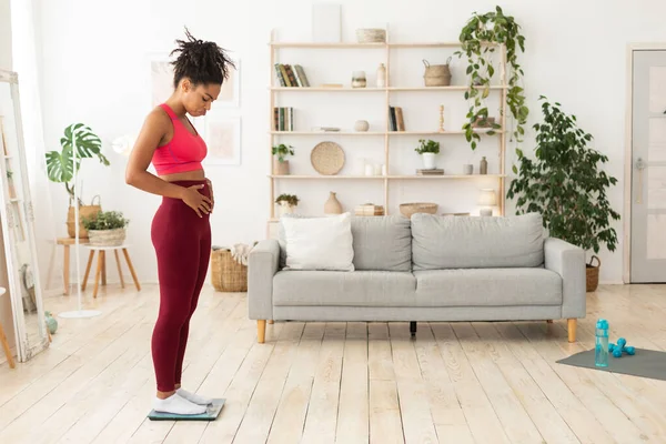 Несчастная афроамериканка, стоящая на весах, прикасаясь к животу в помещении — стоковое фото
