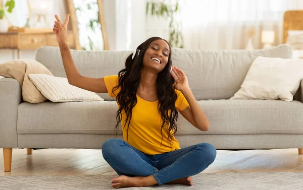 Positive young black woman in headphones listening to music on floor in living room — Stock Photo, Image