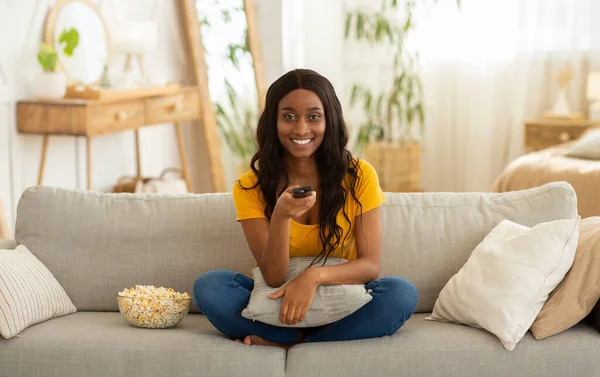 Entretenimiento en casa. Mujer negra sonriente con control remoto comiendo palomitas de maíz y viendo televisión en el sofá en la sala de estar —  Fotos de Stock