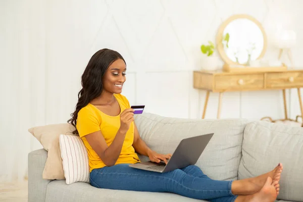 Fare shopping a casa. Attraente signora nera con carta di credito e laptop fare acquisto online sul divano al chiuso — Foto Stock