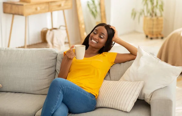 Όμορφη νεαρή μαύρη κυρία αναπαύεται στον καναπέ με ένα φλιτζάνι ζεστό ρόφημα στο σπίτι — Φωτογραφία Αρχείου