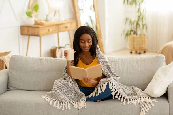 Mujer afroamericana con taza de café leyendo fascinante libro mientras se acurruca bajo tibios cuadros en el sofá en casa — Foto de Stock